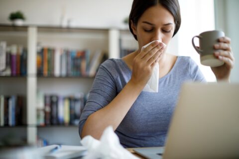 A woman sits at a desk with a laptop, using a tissue due to an allergy in Burbank, CA