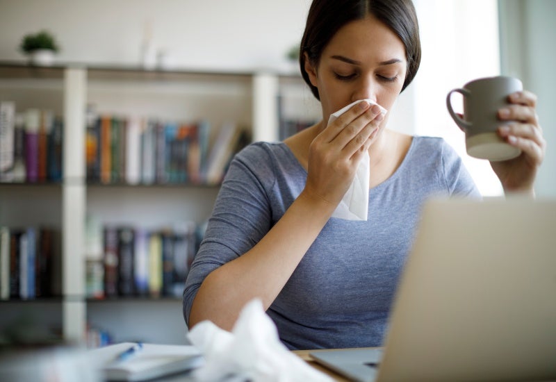 A woman sits at a desk with a laptop, using a tissue due to an allergy in Burbank, CA